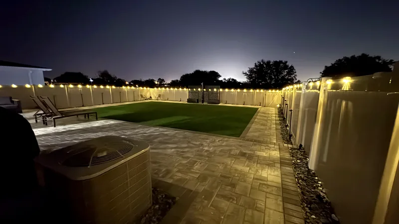 Paver patio and turf lawn at dusk, with lighting running the length of the fence